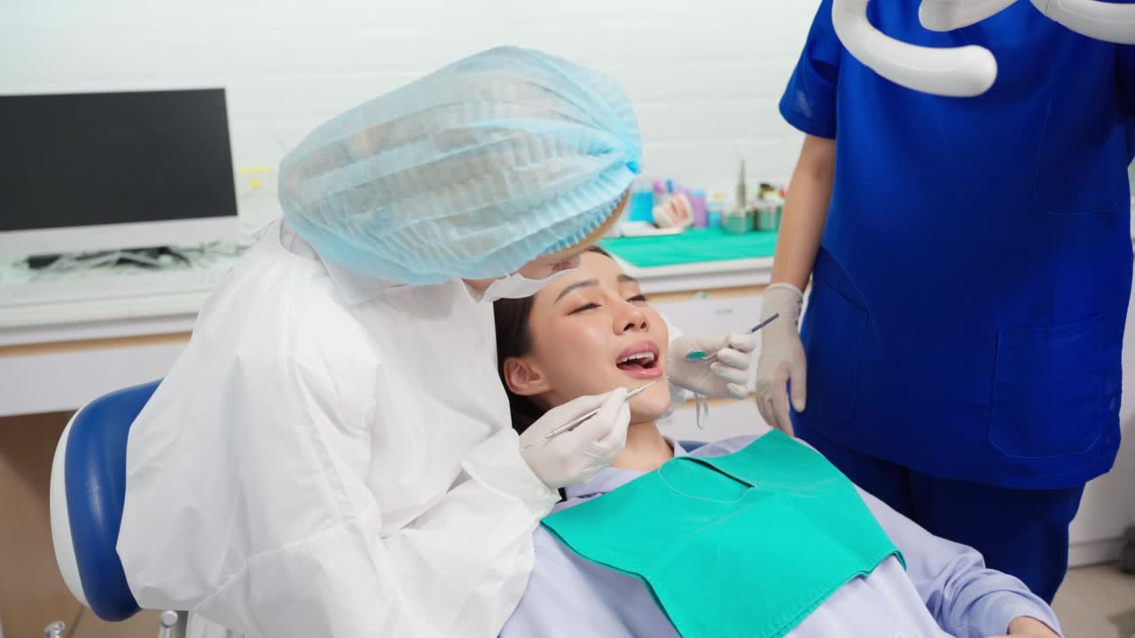 女牙医在牙科诊所为年轻的亚洲女孩检查牙齿。漂亮的女病人躺在牙科椅上，在医院预约手术时接受医生的牙科治疗。视频素材