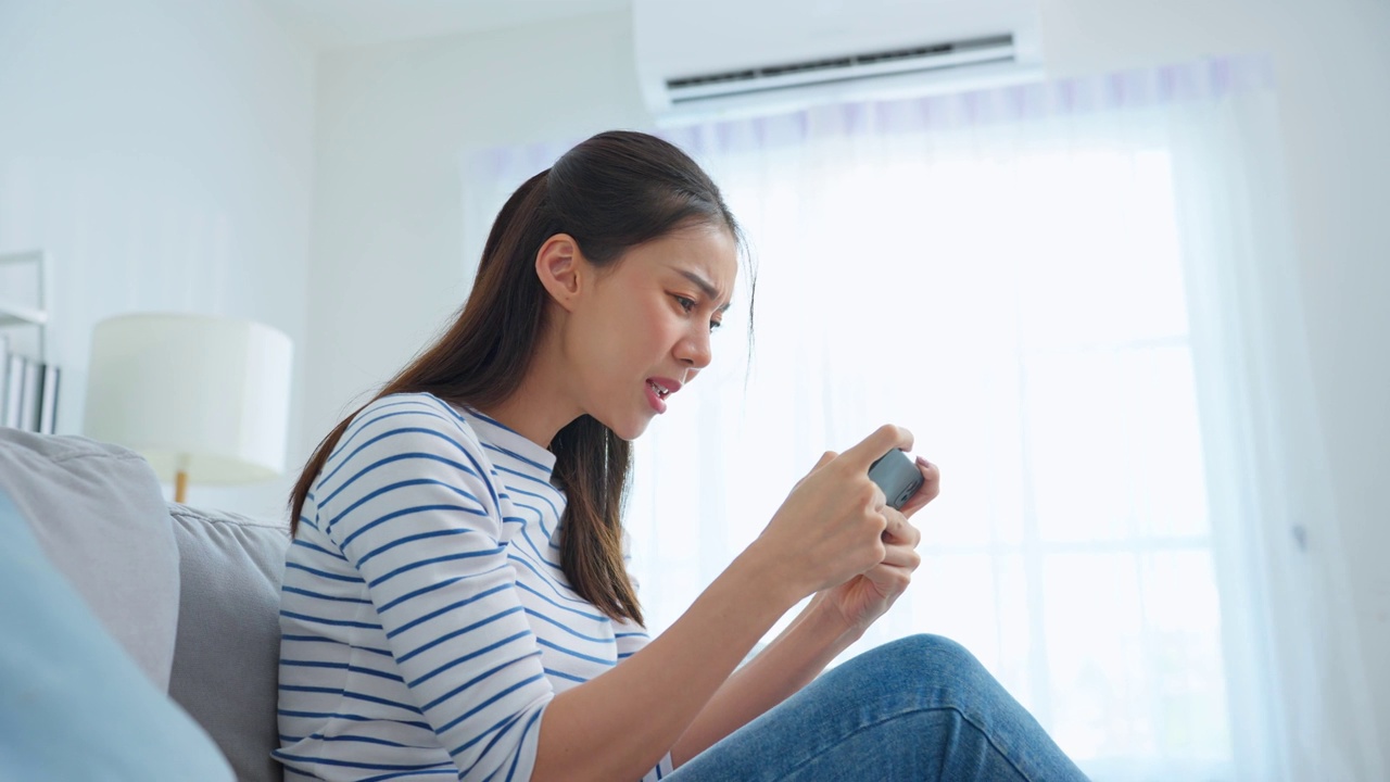 亚洲年轻漂亮的女子在家里玩智能手机游戏。迷人的休闲女孩感到快乐和放松，坐在沙发上玩着玩着手机屏幕播放视频享受胜利在家里视频素材