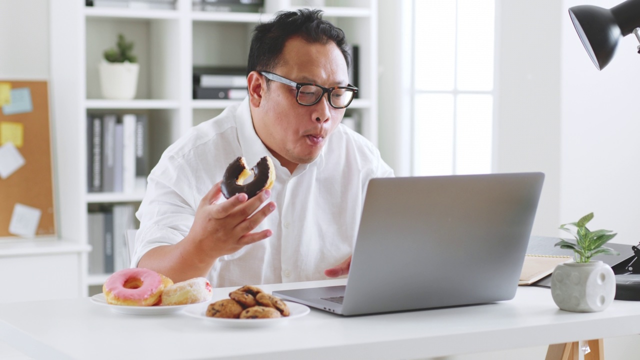 亚洲胖子在用笔记本电脑工作时吃甜甜圈、甜食、垃圾食品，不健康的饮食观念视频素材