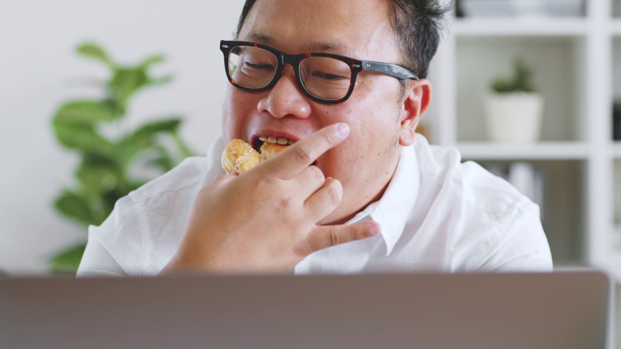 亚洲胖子在用笔记本电脑工作时吃甜甜圈、甜食、垃圾食品，不健康的饮食观念视频素材