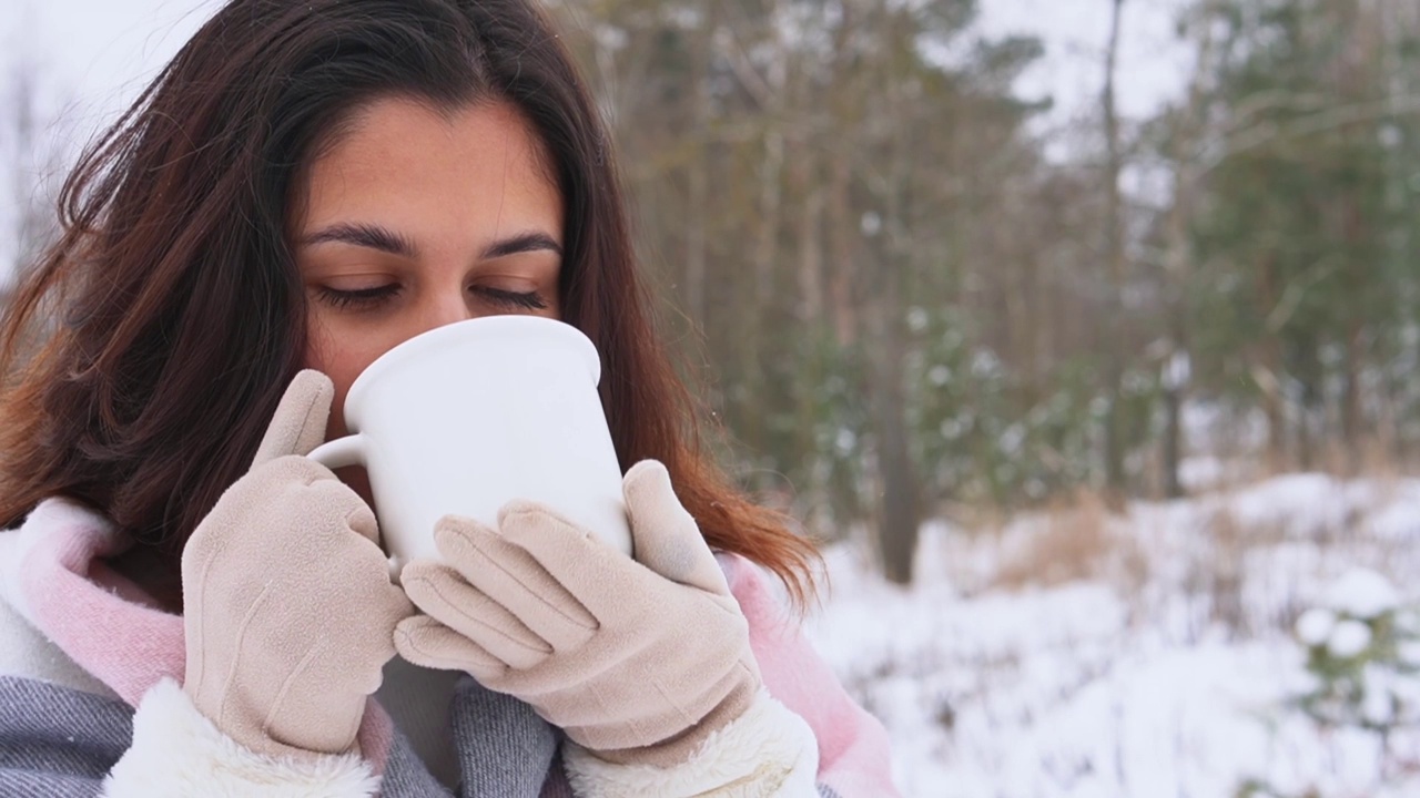 年轻可爱的黑黝黝的黑发女人喝热饮从白色陶瓷杯在积雪的冬天森林视频素材