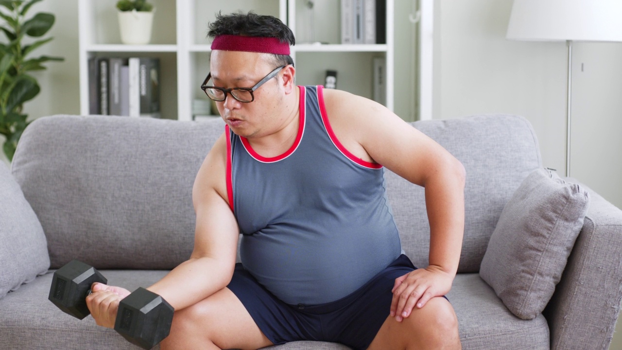 肥胖的中年亚洲男子穿着运动服，在家里用哑铃锻炼视频素材