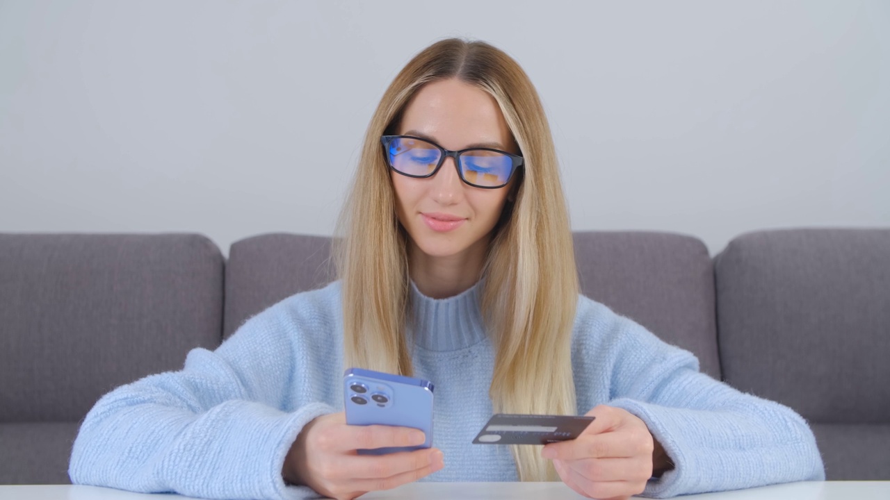 金发女子用信用卡和智能手机在网上购物。年轻女性用银行卡在手机上支付视频素材