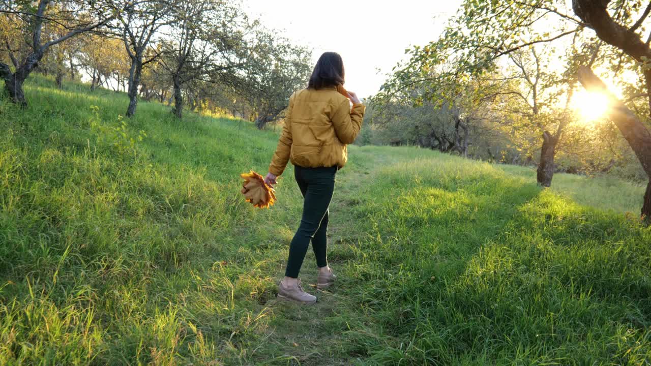 一个穿黄色夹克的褐发女子的后视图。一个女人走在秋天的花园里视频素材