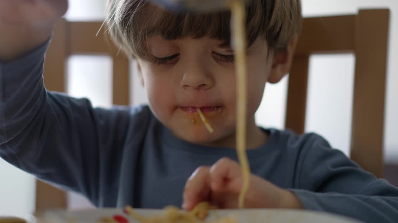 一个有趣的小男孩独自吃意大利面。孩子脏嘴吃用叉子和手面条午餐饭视频素材