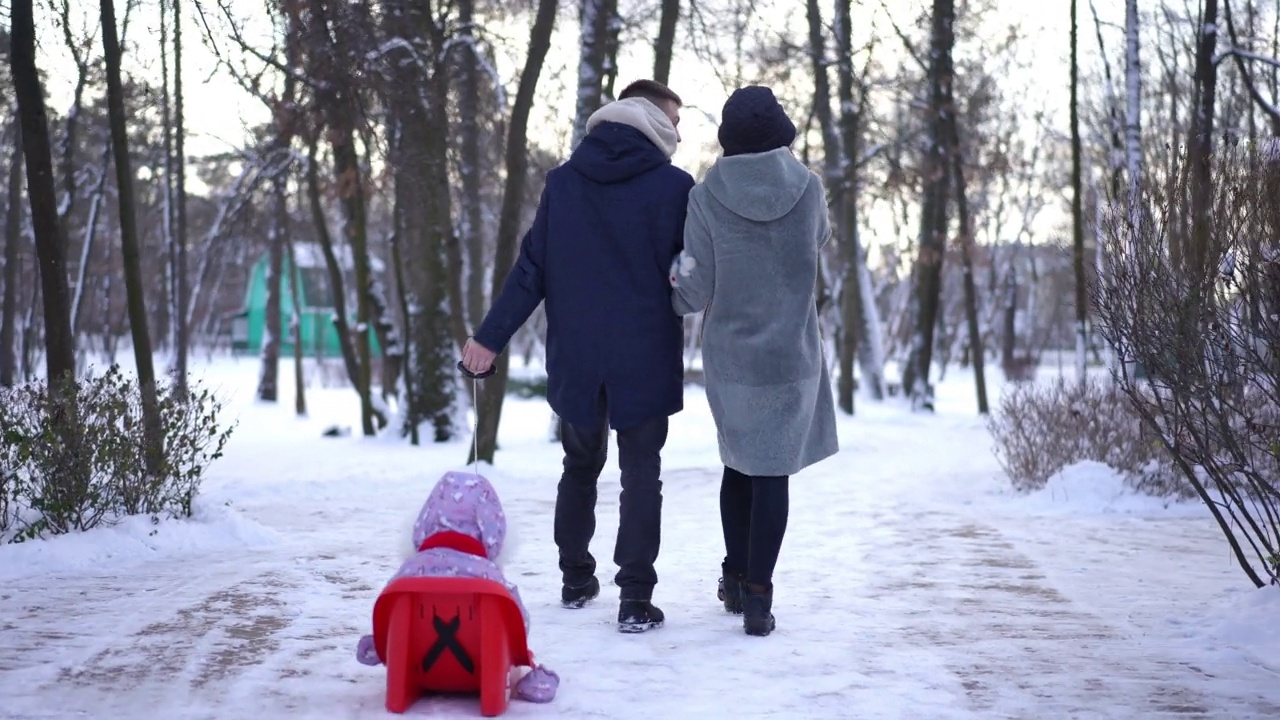 背影快乐的年轻父母在冬季公园里拉着雪橇散步，孩子们在聊天。宽镜头爱的白人男子和妇女享受休闲散步户外与婴儿坐在雪橇。视频素材