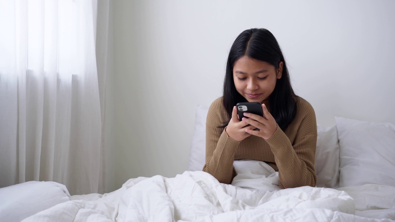 一个坐在床上的女孩喜欢在白天用智能手机发短信或上网聊天视频素材