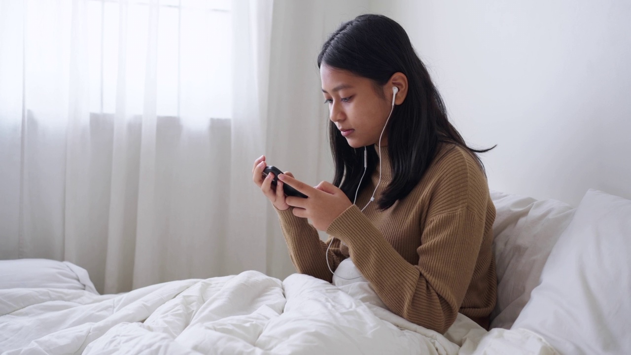 坐在床上的亚洲女孩喜欢在白天用智能手机在线听音乐视频素材