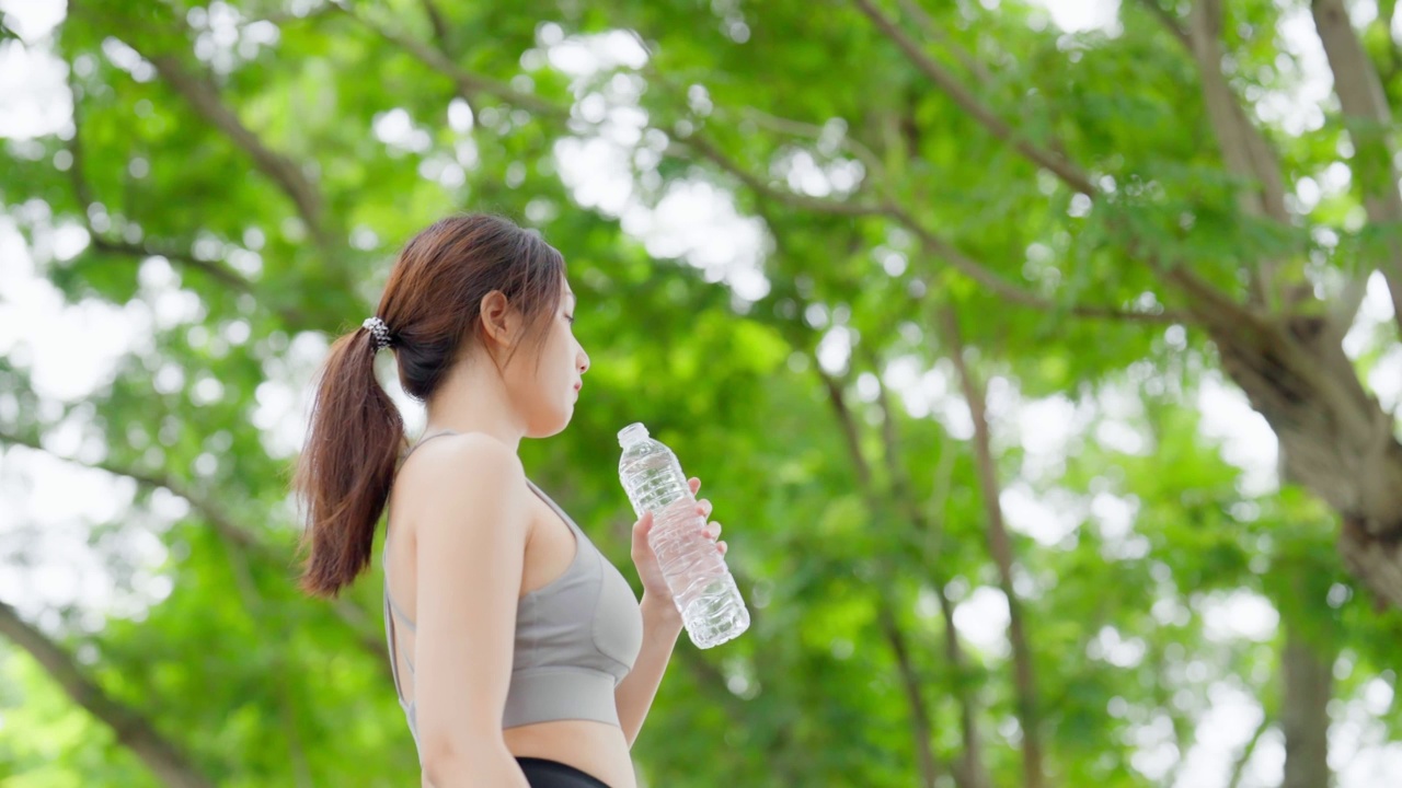 年轻的亚洲妇女在锻炼期间休息时喝瓶子里的水。快乐的青春期女性在公园里锻炼，休息休息，喝点水视频素材