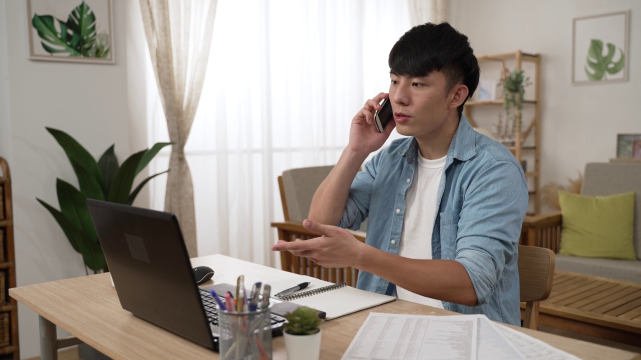 在家办公的亚洲男性在客厅里与业务伙伴讨论财务问题时，一边看电脑一边用手势打电话视频素材