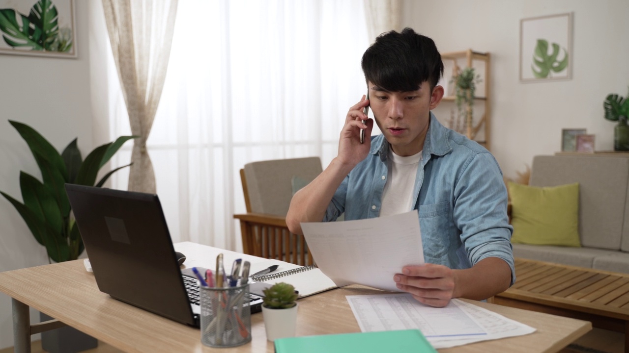 亚洲男性经理接听同事的手机电话，在客厅的电脑上在家工作时查看财务文件视频素材