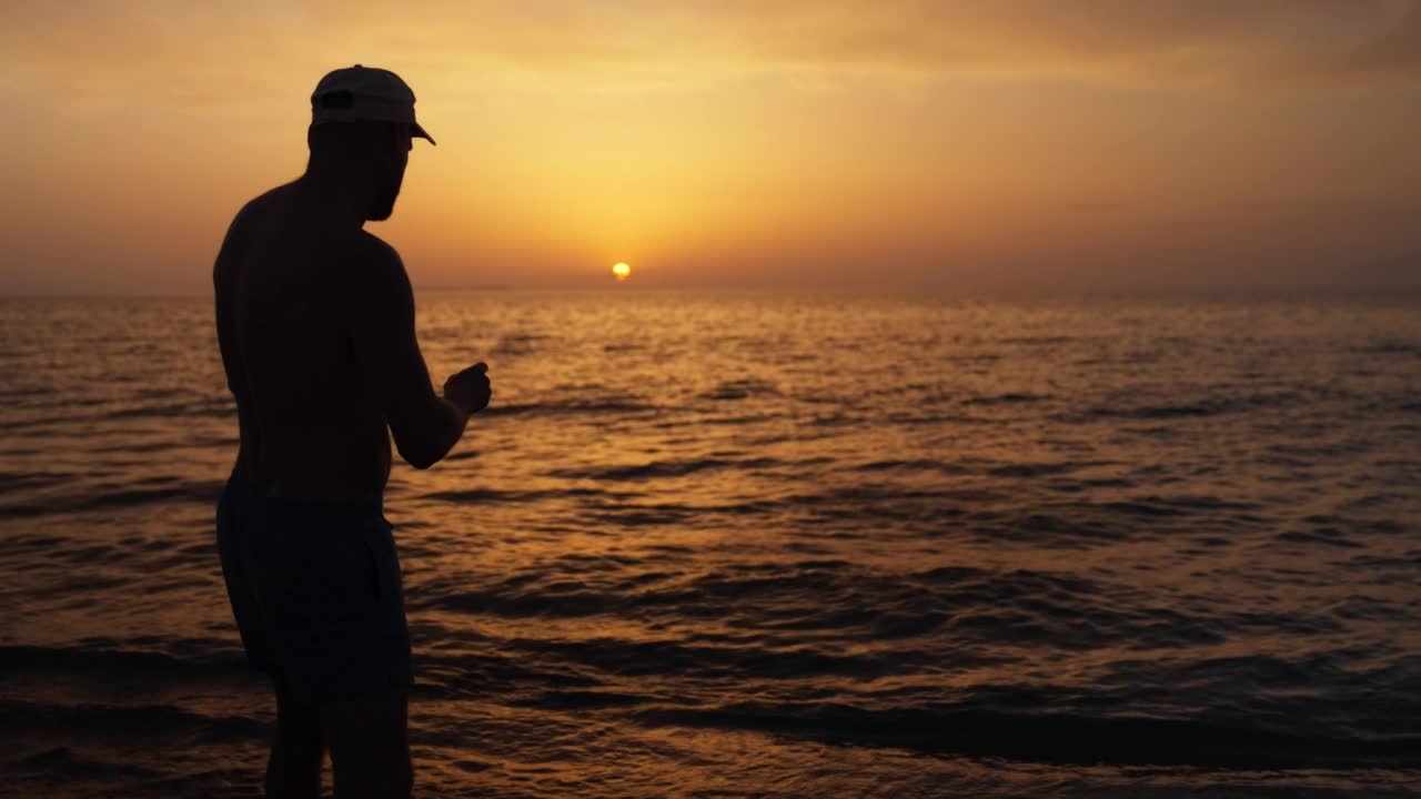 海滩上一个男人的剪影，用慢动作把一块石头扔进海里视频素材