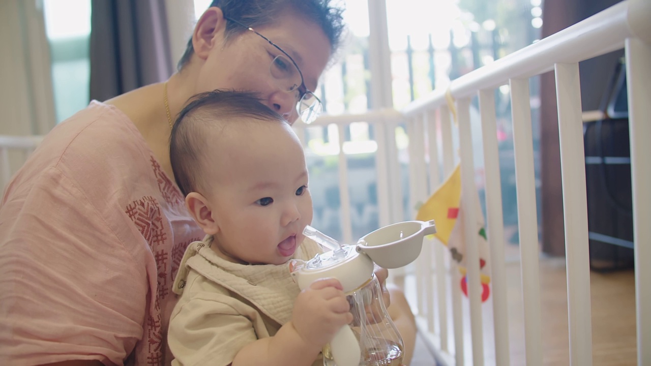 快乐的家庭快乐的资深亚洲妇女奶奶抱着照顾可爱的小婴儿蹒跚学步的孙女和拥抱与爱，而坐在室内房间在家里，婴儿喝水视频素材