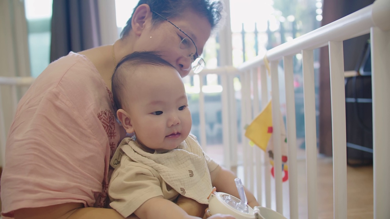 快乐的家庭快乐的资深亚洲妇女奶奶抱着照顾可爱的小婴儿蹒跚学步的孙女和拥抱与爱，而坐在室内房间在家里，婴儿喝水视频素材