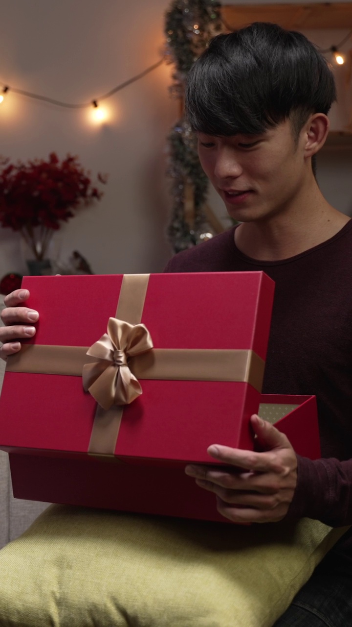 垂直屏幕:有人在家里的客厅靠近圣诞树的地方用礼物给男朋友惊喜。有人坐在室内的沙发上，把大礼物盒送给她的爱人。视频素材