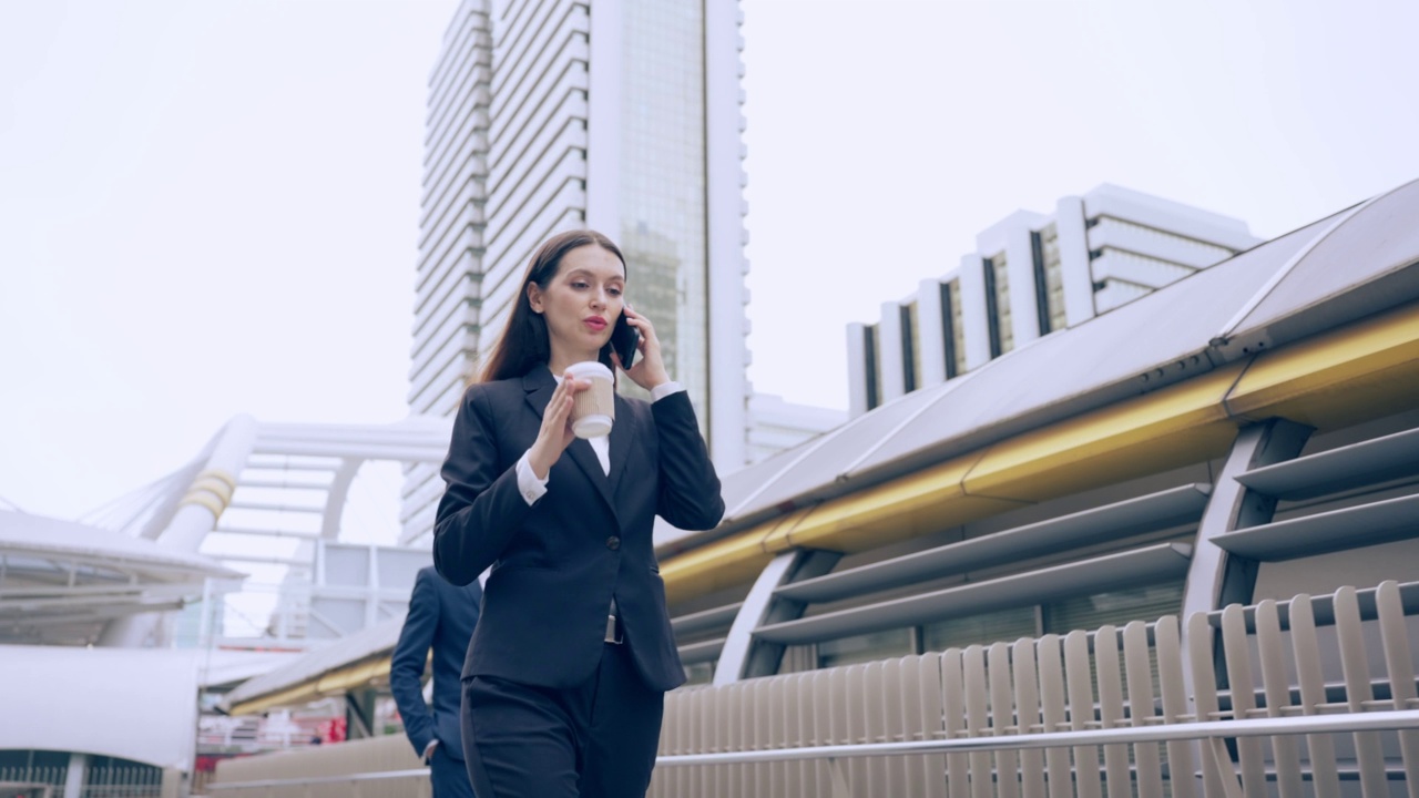 白人年轻女商人在城市里散步时用智能手机说话。迷人美丽的女员工在户外与同事端着咖啡，用手机自信地交流。视频素材