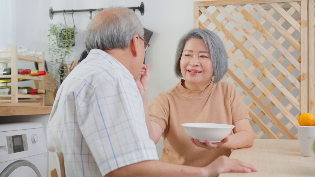 亚洲老年夫妇在家里的厨房里一起吃沙拉。幸福的家庭，有魅力的年长成熟的夫妻坐在餐桌上享受退休生活，午餐吃健康的食物在家里。视频素材