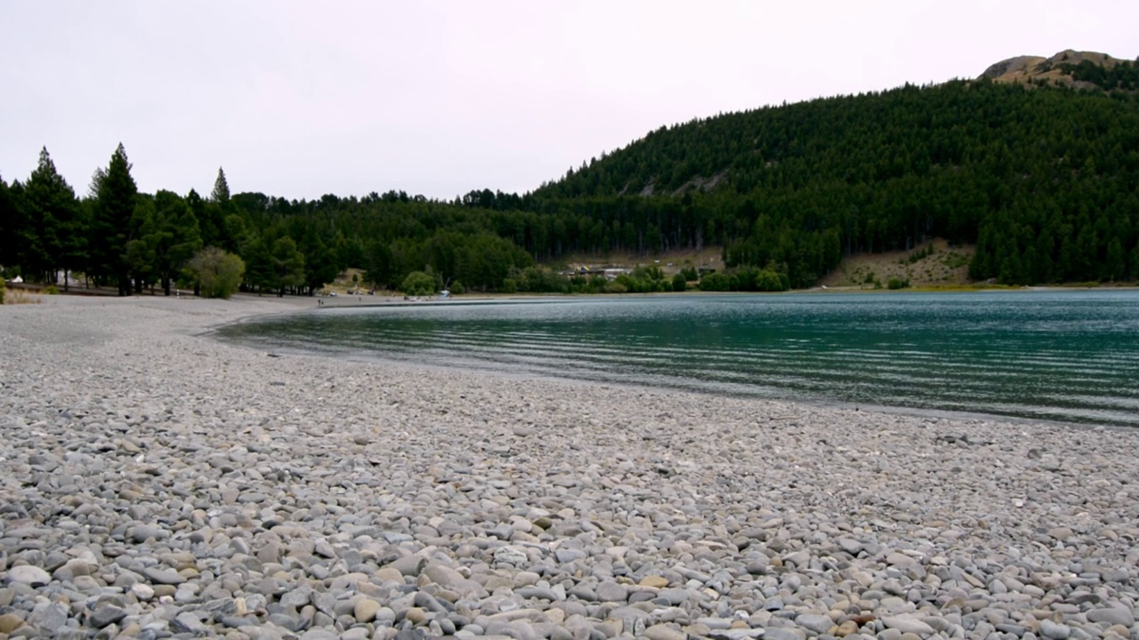 Tekapo湖的波浪冲刷着南岛远处的岩石河岸和森林覆盖的小山视频素材