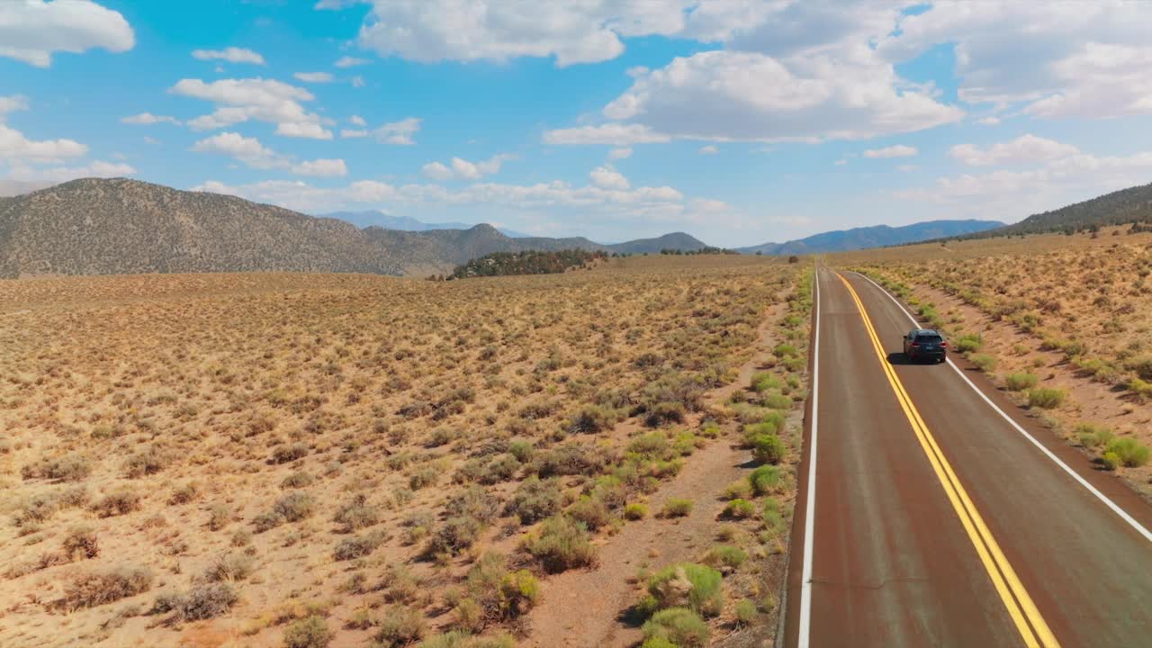 汽车行驶在双车道的道路上，沿着干燥的沙地景观。光秃秃的山在蓝色的天空和蓬松的云的背景。视频素材