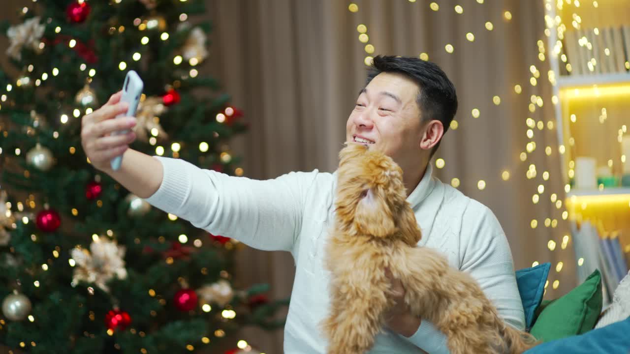 一名亚洲男子带着宠物小狗坐在家里用智能手机通过视频电话问候新年快乐、圣诞快乐。视频素材