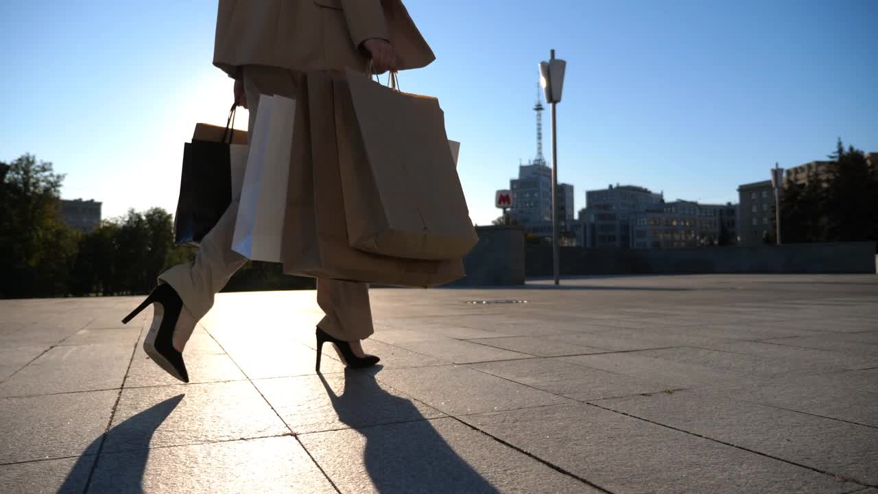 年轻女子拿着购物袋走在人行道上购物后。穿着西装的优雅女孩背着纸袋走在阳光明媚的城市街道上。销售和折扣的概念。特写慢动作视频素材