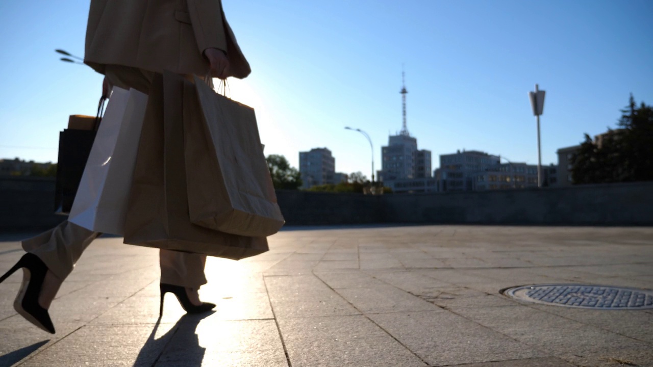 年轻女子拿着购物袋走在人行道上购物后。穿着西装的优雅女孩背着纸袋走在阳光明媚的城市街道上。销售和折扣的概念。特写慢动作视频素材