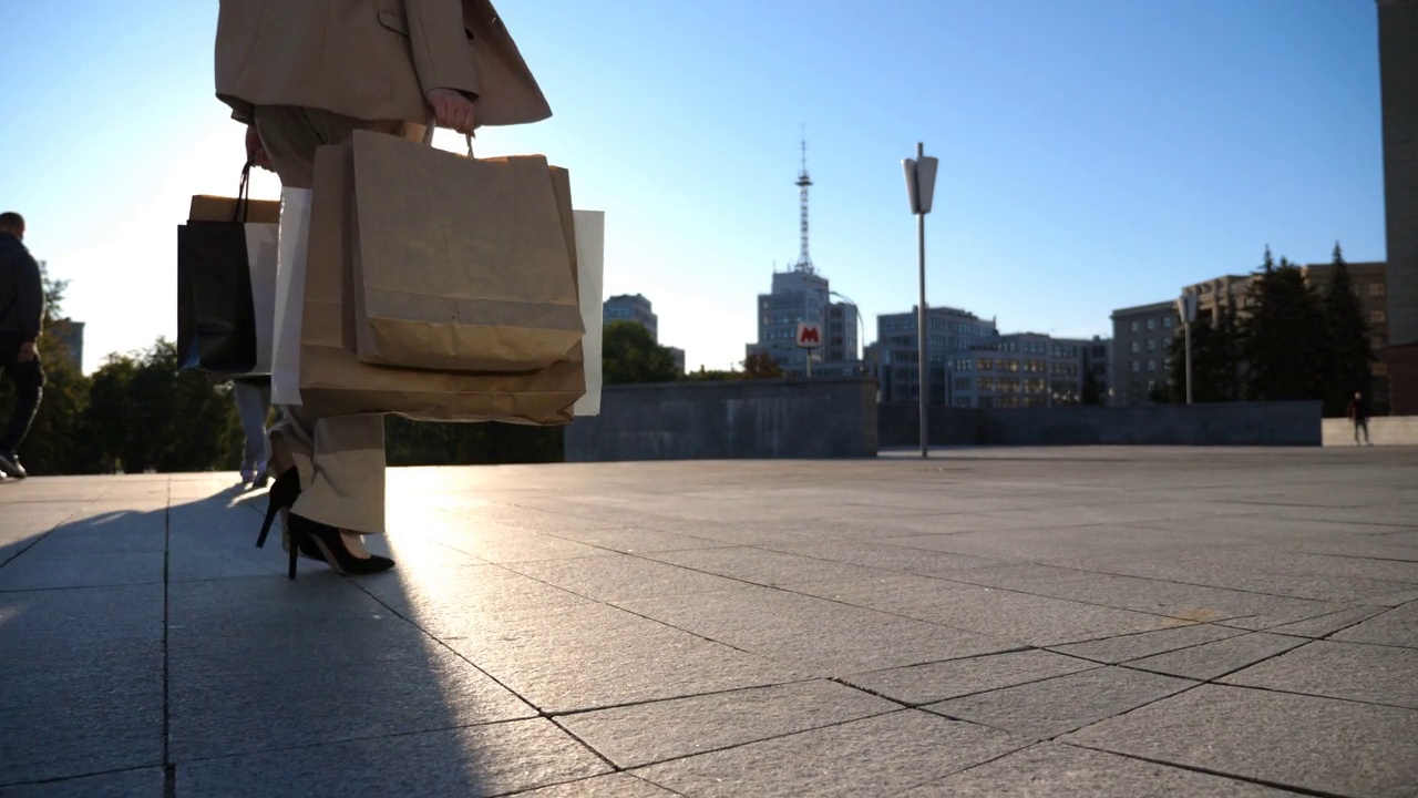 年轻女子拿着购物袋走在人行道上购物后。穿着西装的优雅女孩背着纸袋走在阳光明媚的城市街道上。销售和折扣的概念。特写慢动作视频素材