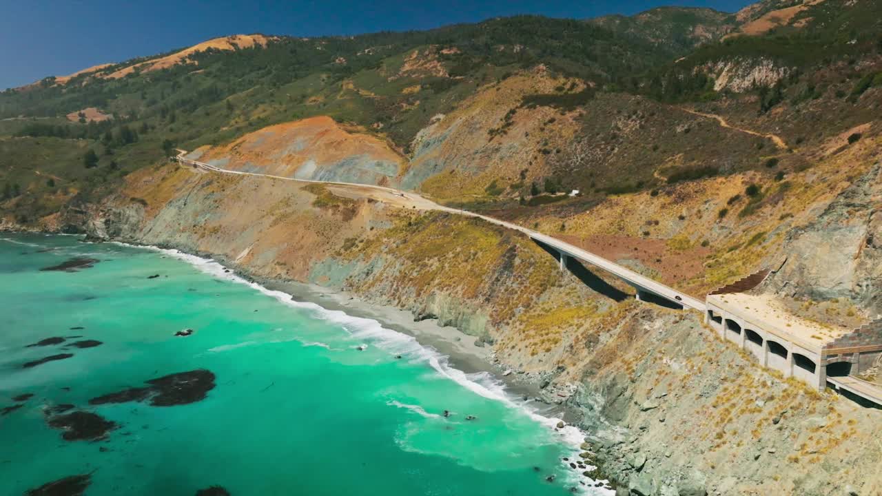 令人惊叹的碧蓝的太平洋海水和泡沫波到达岩石海岸。在炎炎夏日，海岸线上的山脉有高速公路和桥梁。前视图。视频素材