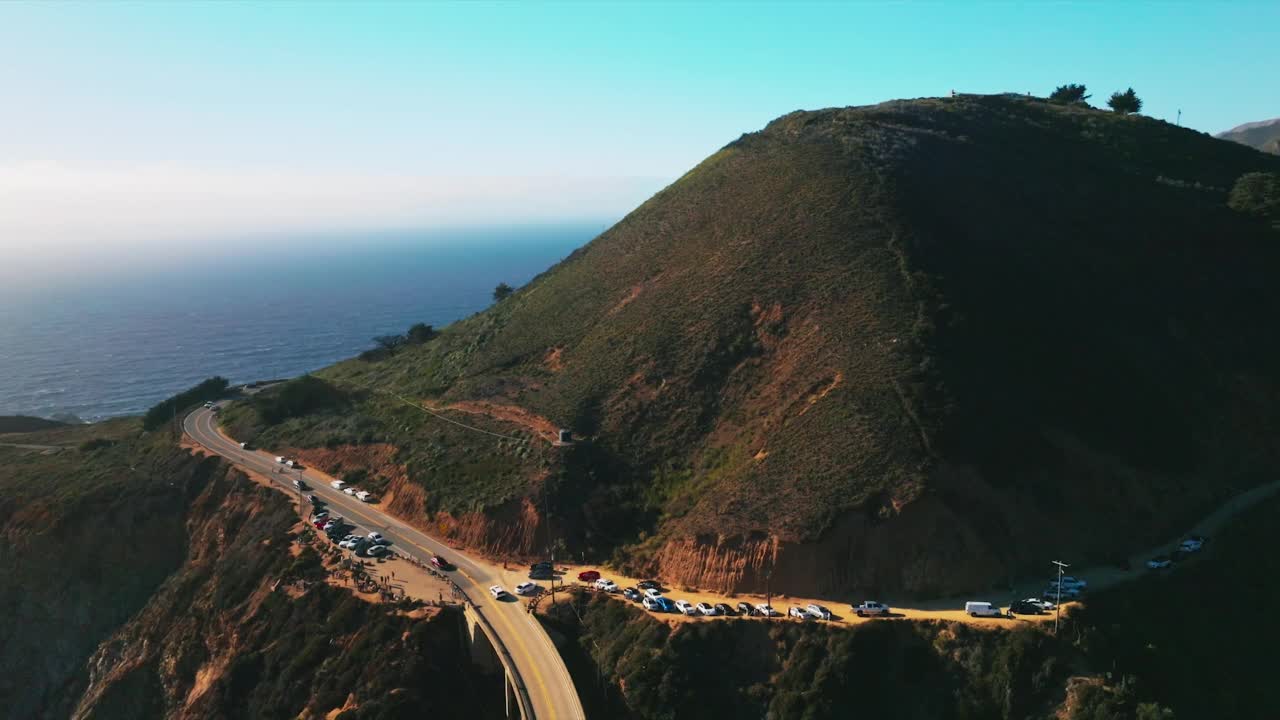 沿着长满绿草的山坡上升。山脚下有高速公路，山顶有房子。海洋与天空融为一体的壮丽景色。视频素材