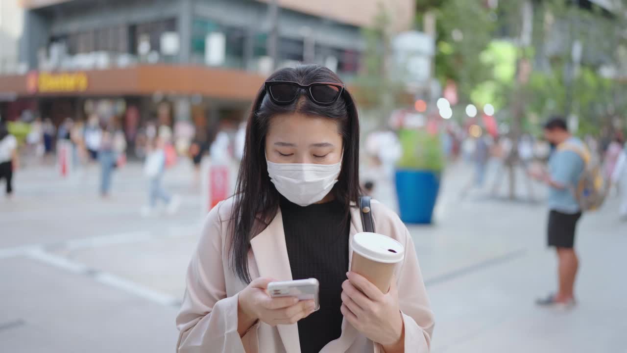 亚洲妇女戴着口罩，用着智能手机，端着一杯咖啡走在城市街道上视频素材