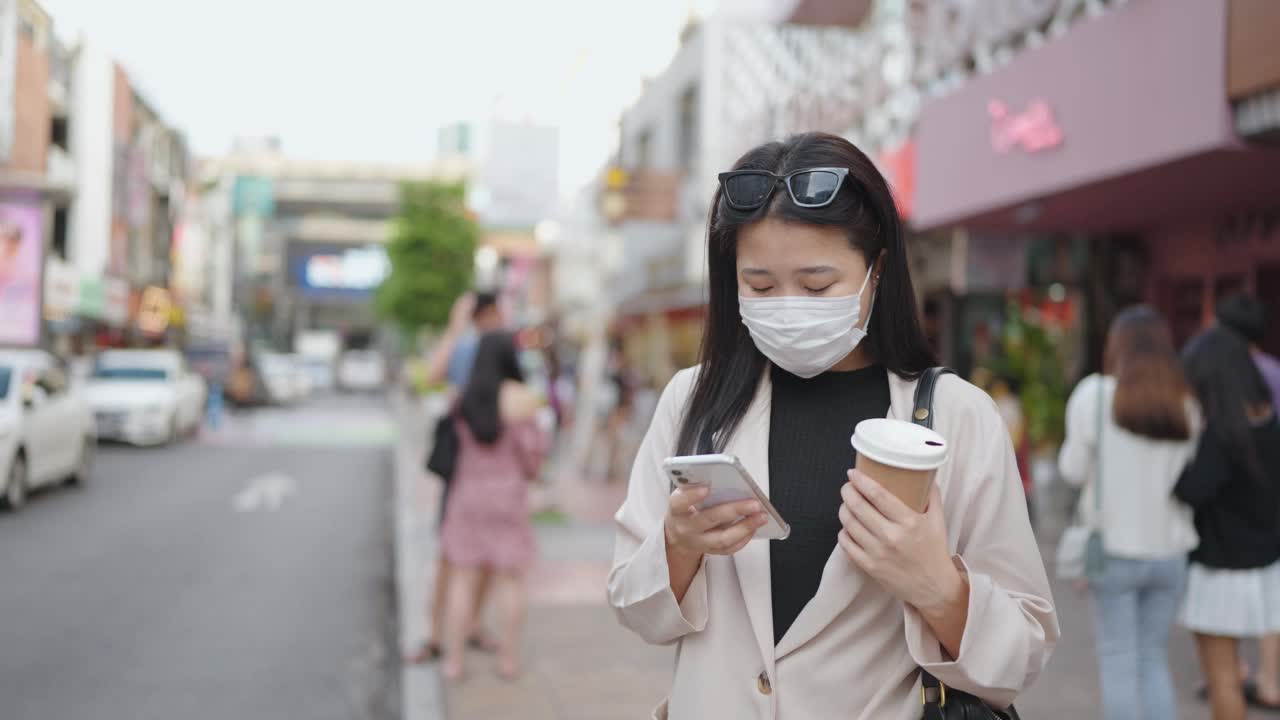 亚洲妇女戴着口罩，用着智能手机，端着一杯咖啡走在城市街道上视频素材