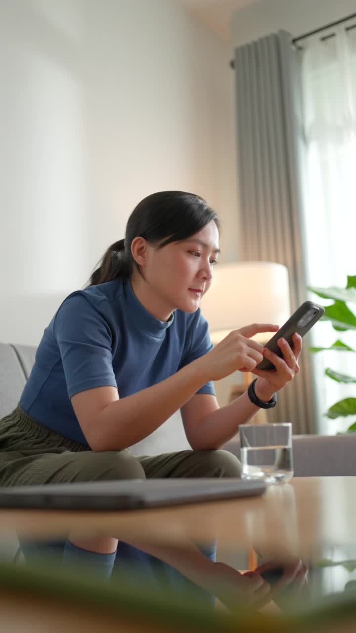 亚洲妇女快乐地微笑着坐在沙发上拿着杯子，在家里使用智能手机。视频素材