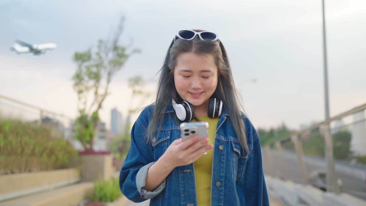 亚洲女性旅行者微笑着用智能手机走路。背景天空和飞机视频素材