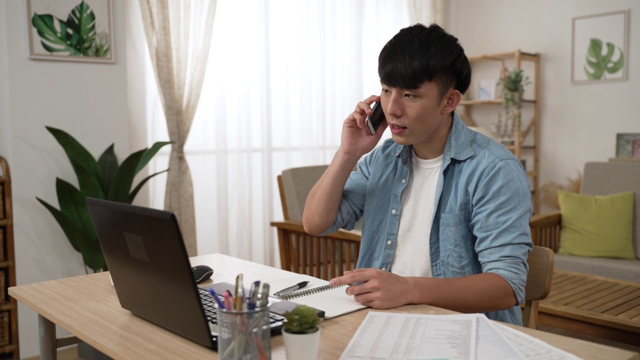 在家办公的亚洲男性在客厅里与业务伙伴讨论财务问题时，一边看电脑一边用手势打电话视频素材