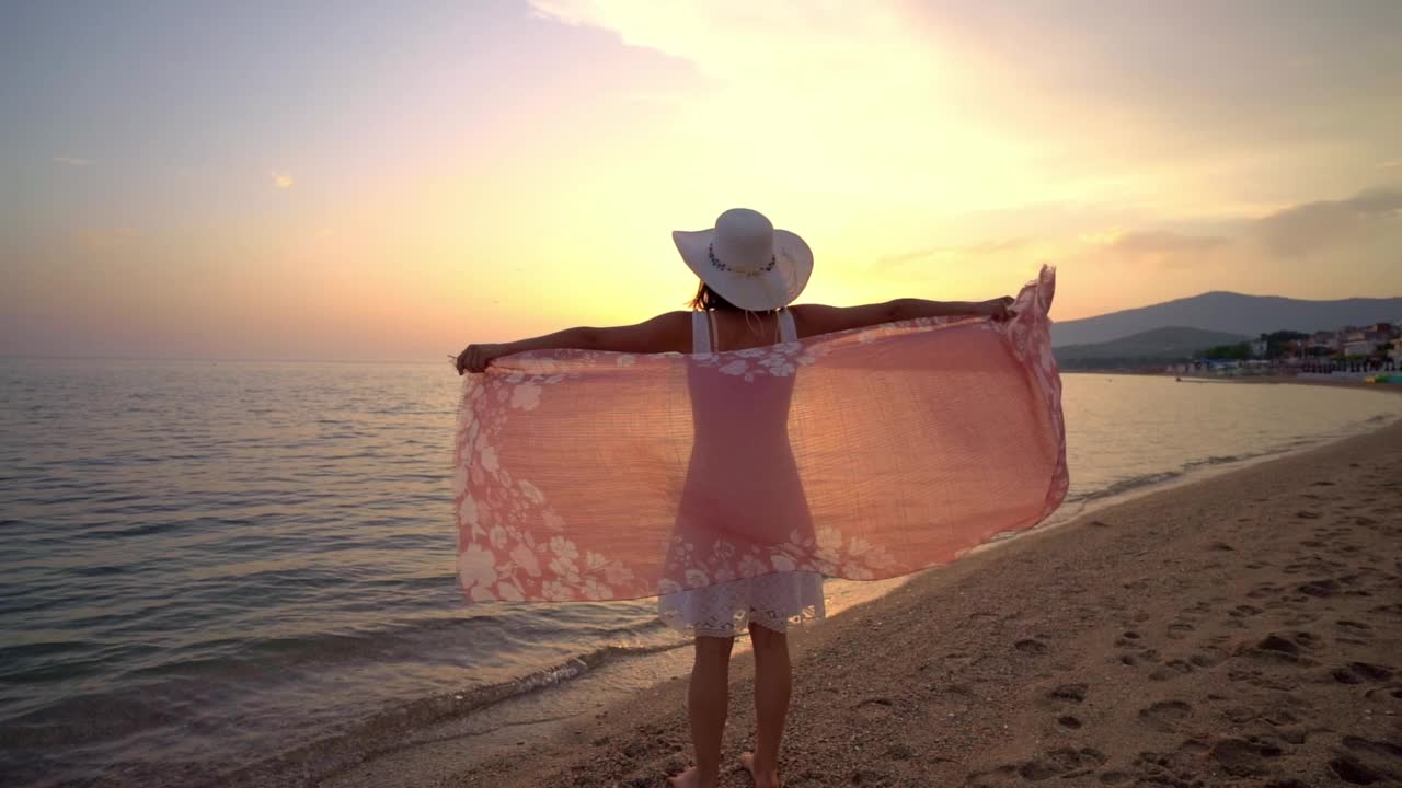 一个女人在海边欣赏日落，她伸出双臂，拿着围巾视频素材