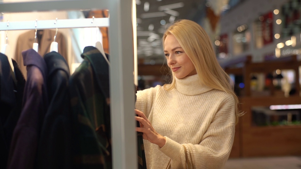 迷人的微笑着金发年轻女子在服装店选择外套，站在女性服装陈列柜附近。视频素材