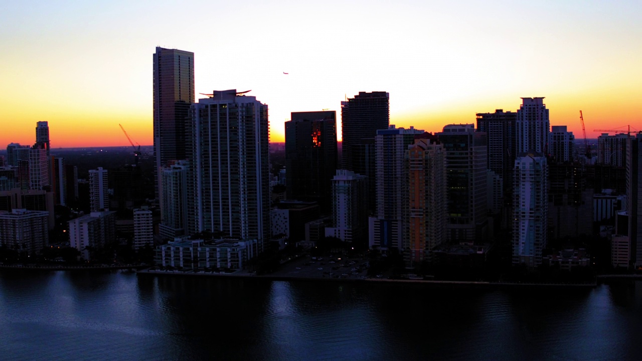 空中拍摄的飞机飞过现代化的城市建筑，无人机在夕阳下的海浪上移动-迈阿密，佛罗里达州视频素材