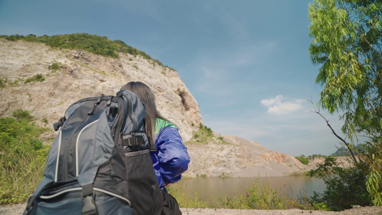 穿着蓝色雨衣背着背包的女性徒步旅行者在山上行走。迷人快乐的年轻女子背着背包徒步旅行视频素材