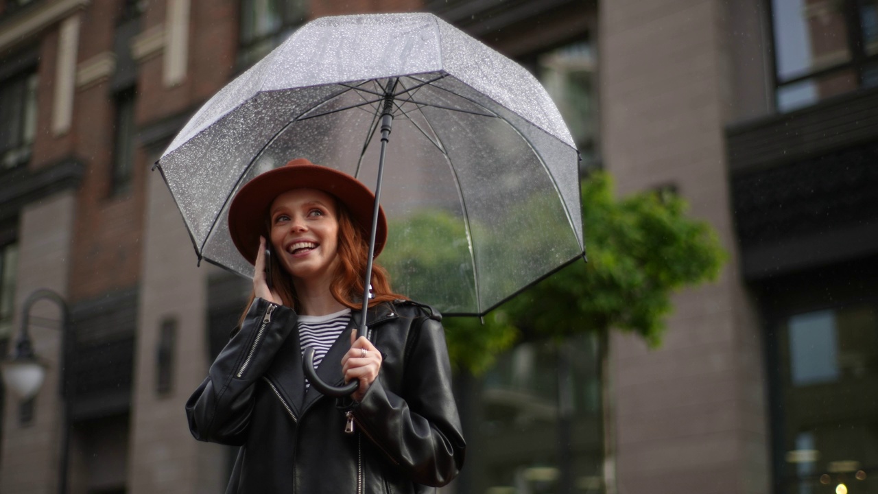 特写脸快乐美丽的女人戴着帽子和透明伞在雨中在城市街道上打电话。视频素材