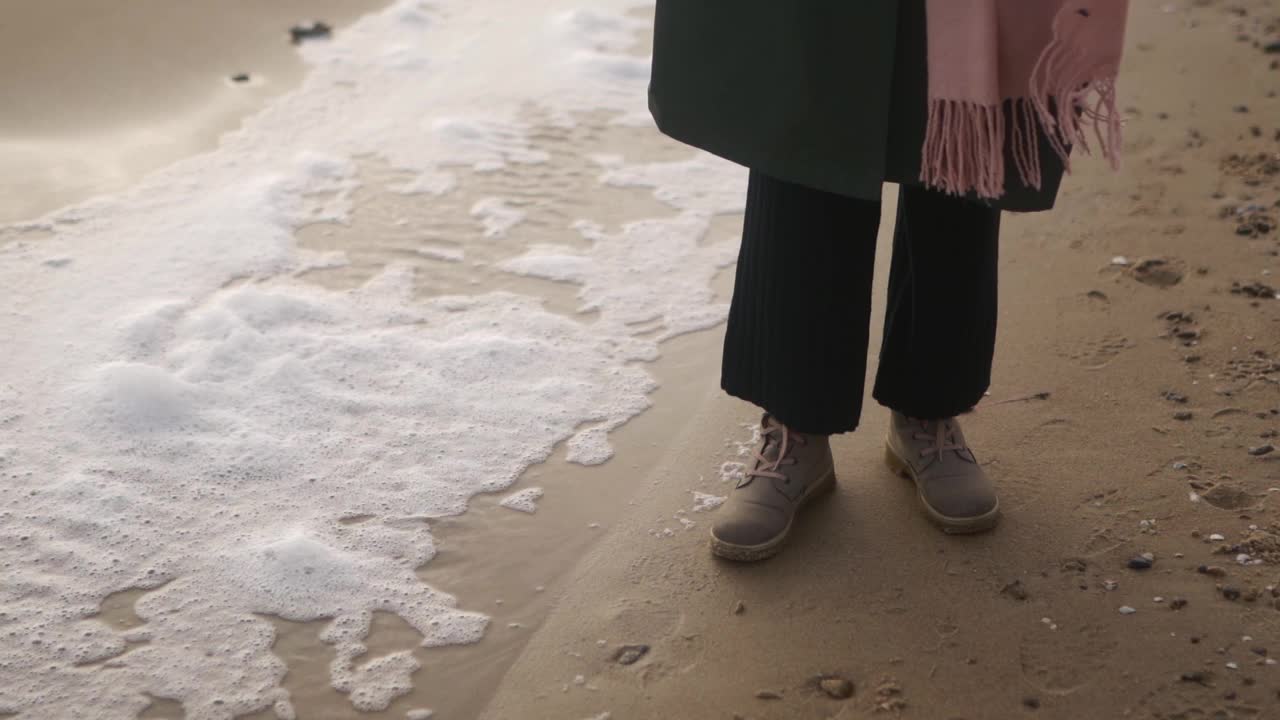 在慢动作的沙滩上，女人穿着靴子站在风吹起的海水泡沫旁。在狂风暴雨的日子里，女性脚上的防水鞋被海浪的泡沫流片冲上岸。低角度。视频素材