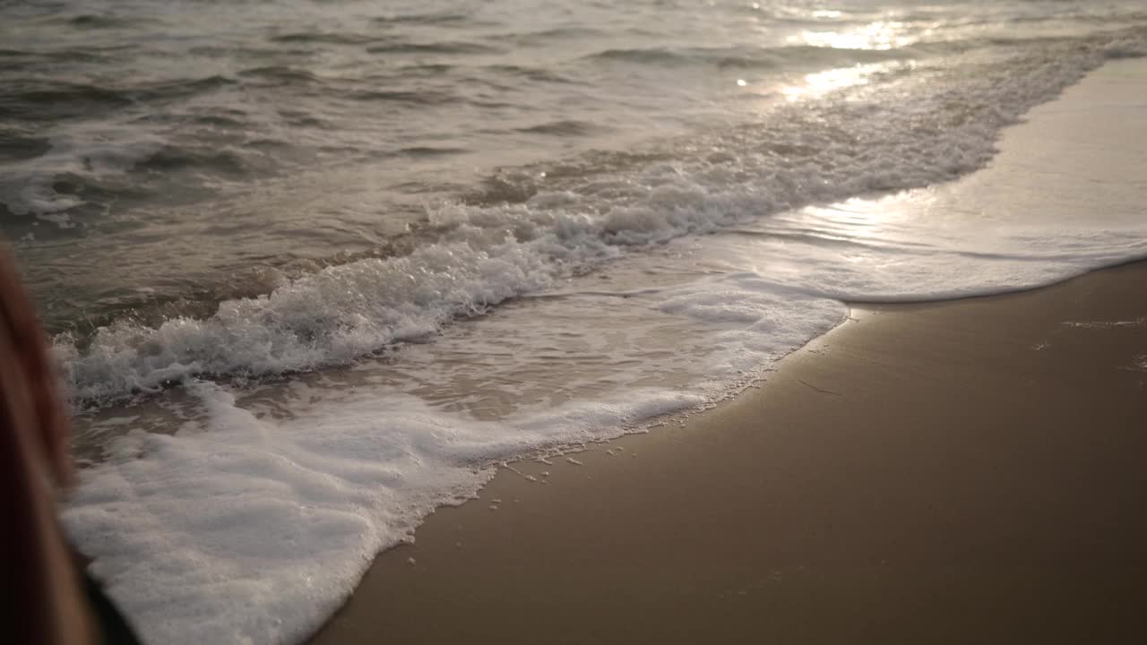 侧视图女性脚在靴子沿着海浪泡沫在沙滩上行走。穿着绿色外套的女人沿着海岸线上的潮汐前进。倾斜建立梦幻的电影去个性化的镜头。视频素材