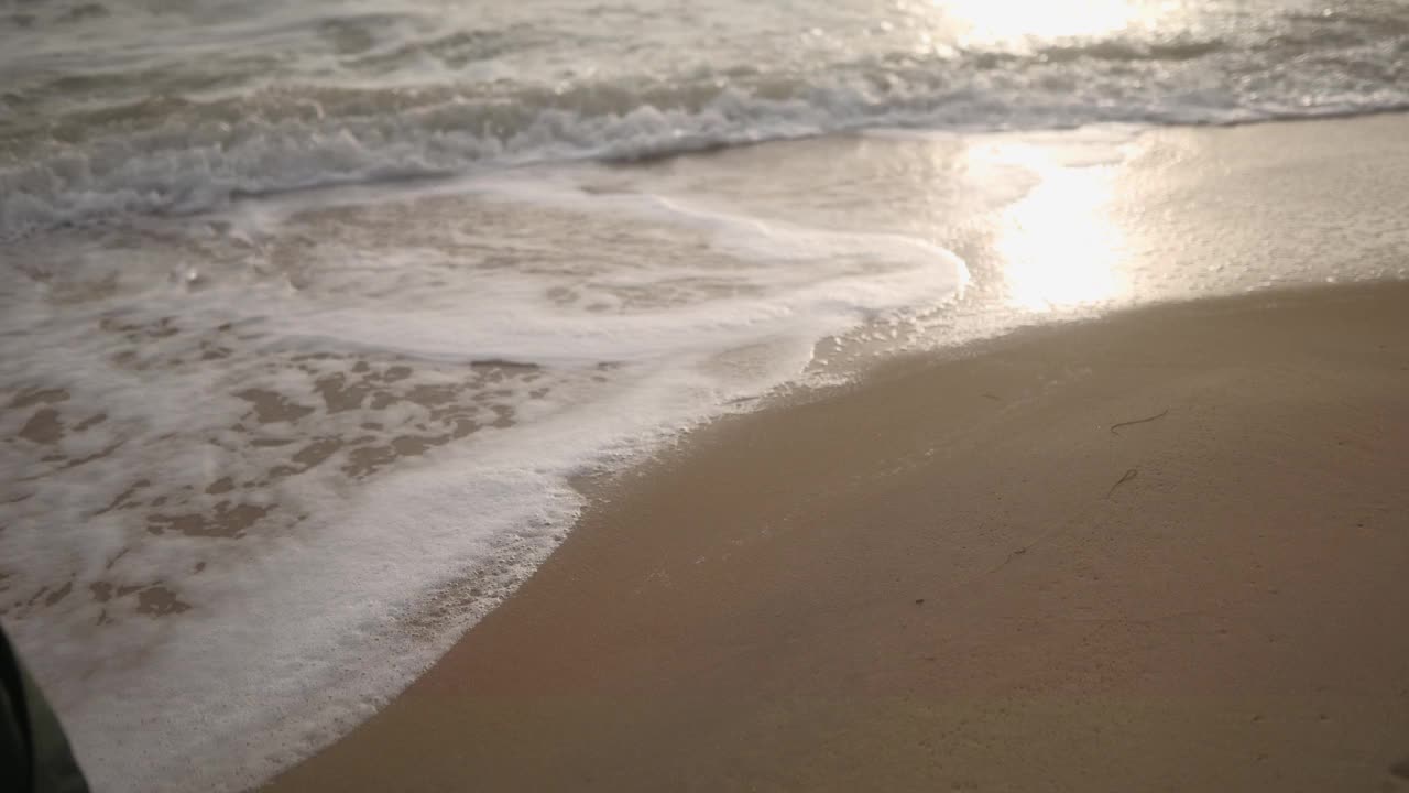 侧视图女性脚在靴子沿着海浪泡沫在沙滩上行走。穿着绿色外套的女人沿着海岸线上的潮汐前进。倾斜建立梦幻的电影去个性化的镜头。视频素材