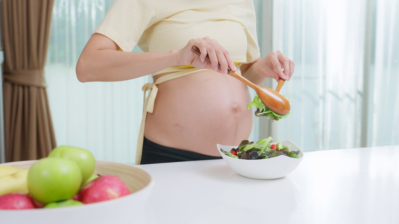 亚洲美丽的孕妇在厨房做饭健康的食物在家里。漂亮的怀孕女孩站在室内的厨房里感到快乐，享受用碗做绿色蔬菜沙拉在家里的健康视频素材