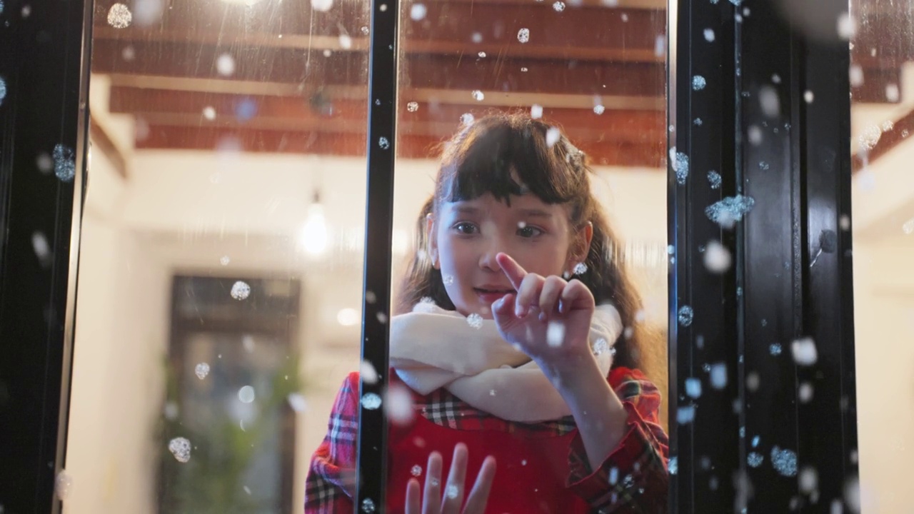 可爱的孩子透过窗户欣赏着初雪。小女孩坐在家里的客厅里，看着外面飘落的雪花享受着与家人一起庆祝圣诞节、感恩节。视频素材