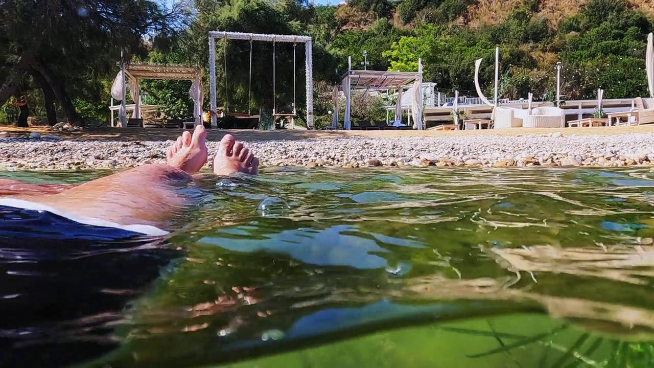 半水下的观点，男性的腿和脚放松，同时漂浮在海水与藻类和事件设置在沙滩上摇摆的背景。慢动作的暑假概念视频素材