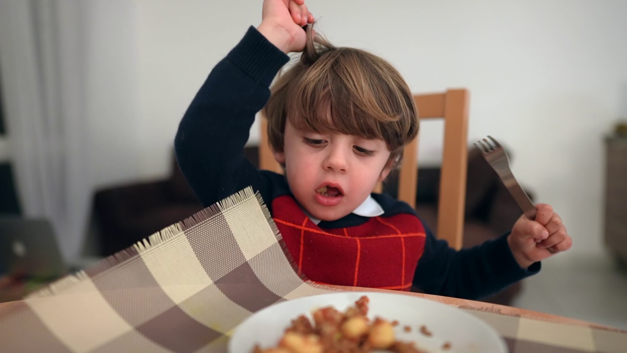 快乐的小孩用叉子自己吃食物。一个小男孩在冬天穿着毛衣吃晚饭视频素材