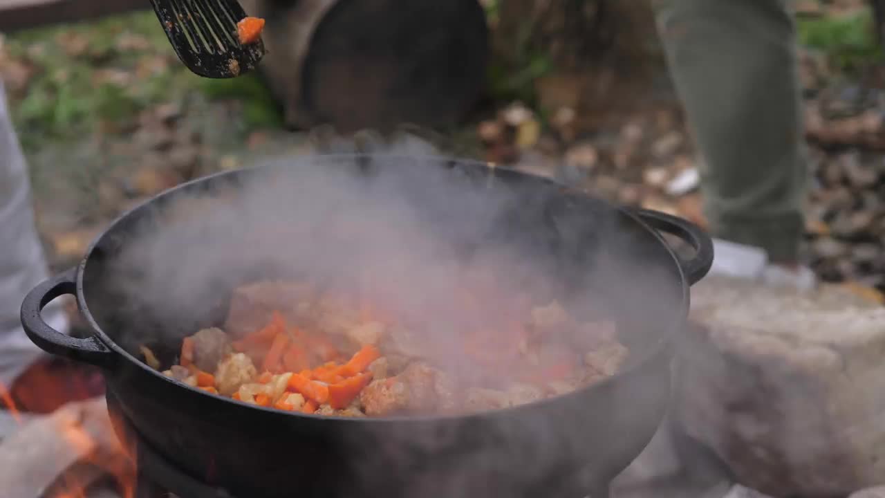 一名男子把用来做抓饭的米饭倒进肉里，把蔬菜切成块放在一个铸铁大锅里。在户外露营时烹饪食物。在篝火上做饭，野餐。明火准备工作视频素材