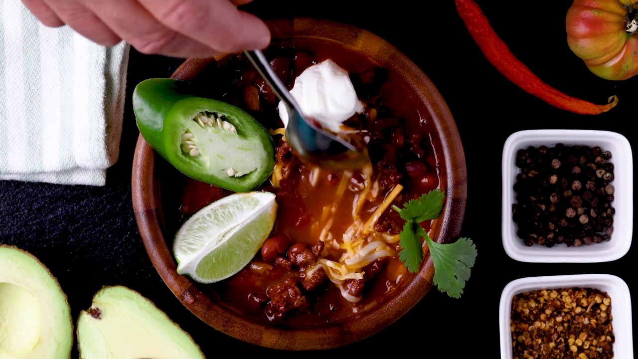 Chili garnished with lime, green jalapeño pepper, sour cream, cilantro and shredded cheese. Spices and vegetables spilled around it.视频素材