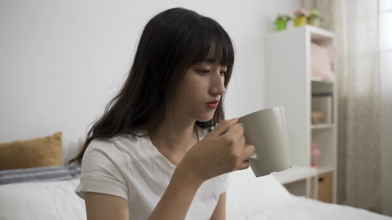 快乐的亚洲女孩闻着茶杯里的早茶，点点头，对家里卧室里的好品质感到满足。视频素材