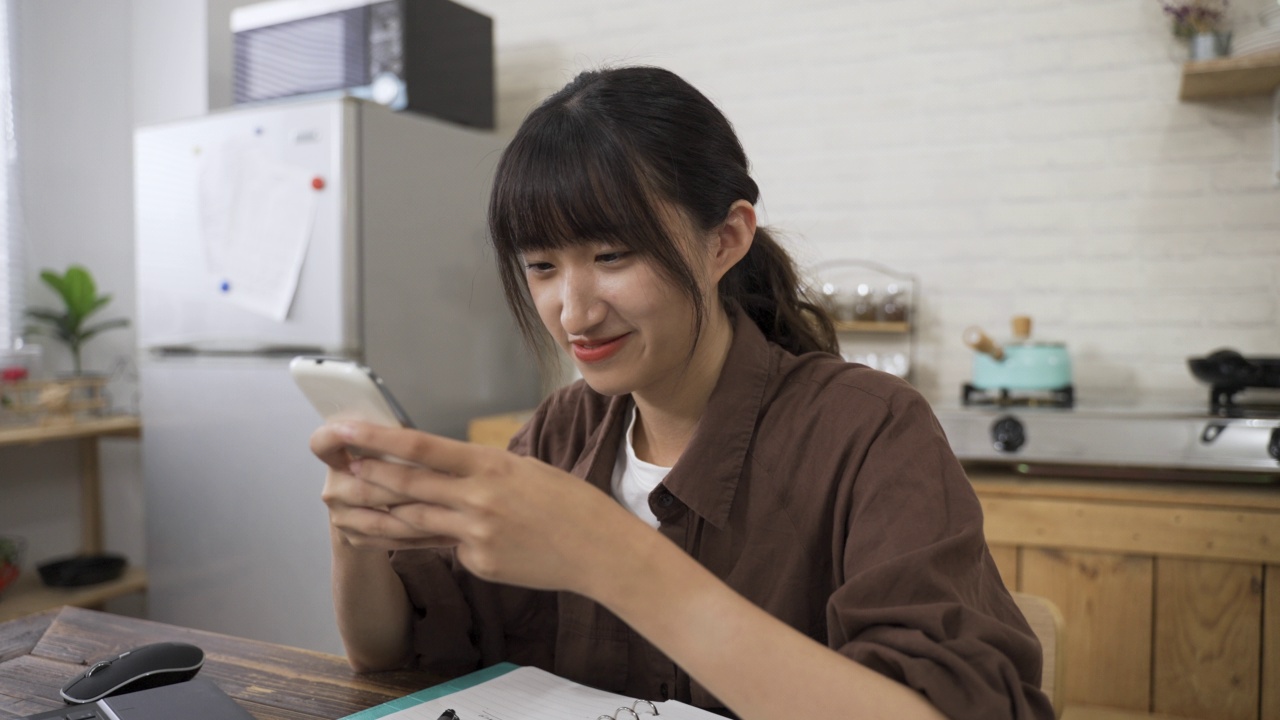 在明亮的饭厅里，一位面带微笑的台湾年轻女士享受着通过即时通讯应用聊天的手机特写镜头视频素材